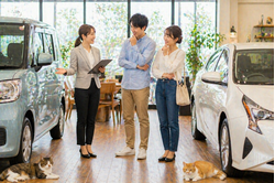 明るい中古車販売店の店内で軽自動車と普通車の間に立ちながらスタッフが男女のお客様に違いを説明し比較しながら悩んでいる相談シーンと足元でくつろぐ猫たち