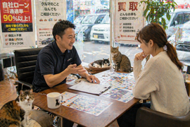 明るい店舗内のテーブルで単独の女性客が少し悩みながらスタッフの説明を受け安心していく相談シーンと足元やテーブル周りでさりげなく見守る猫たち