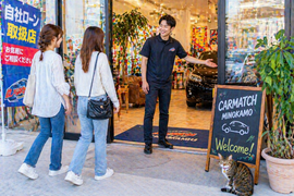 店舗入口でスタッフが笑顔で女性二人組のお客様を店内へ案内している自然な来店シーンと足元でくつろぐ猫