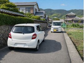 淡路島の細い生活道路で、白いコンパクトカーと軽自動車が対向してすれ違う様子。狭い道での運転や車選びをイメージした風景。
