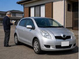 男性が自宅前で中古コンパクトカーと書類を確認している。年金受給者の車購入・自社ローン相談のイメージ。