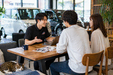明るいショールームのテーブルでスタッフが女性二人のお客様と車の購入や売却について丁寧に相談している自然な商談風景と足元でくつろぐ猫