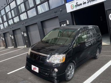 黒のトヨタ ノア（AZR60G）が店舗前に停車している外観写真。両側スライドドアのミニバンで、街中でも扱いやすいサイズ感のファミリー向け車両。