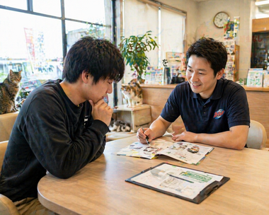 明るい店舗内のテーブルで若い男性が少し不安な表情から安心した様子に変わりながらスタッフと車の購入や売却について相談している商談風景